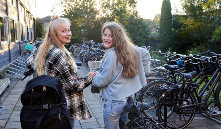 Decoratieve foto van twee leerlingen die op het punt staan weg te fietsen van school. Ze kijken om en lachen naar de camera.