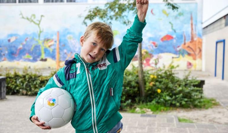 Decoratieve foto van een jongen op het schoolplein. Hij houdt met de ene arm een voetbal van Gezonde School vast. De andere arm steekt hij juichend in lucht.